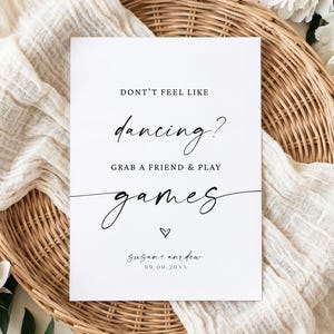 May include: A white rectangular sign with black text that reads: DONT'T FEEL LIKE dancing? GRAB A FRIEND & PLAY games. Below the text is a small heart symbol, the names susan + andrew, and the date 09.09.20XX in a clean, minimalist font style.