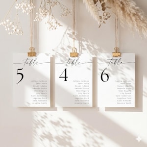 May include: Three white rectangular table seating cards hanging from twine with gold metal clips. Each card features the word Table in black script, a large black number 5, 4, or 6, and a list of names including Ashley Jackson and Jane Jules.