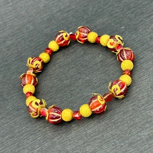 May include: A red and gold beaded bracelet with a textured finish. The beads are round and have a swirl design.