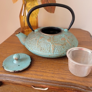 May include: A teal blue cast iron teapot with a black handle and a matching lid. The teapot has a floral design and is sitting on a wooden table with a silver tea strainer.