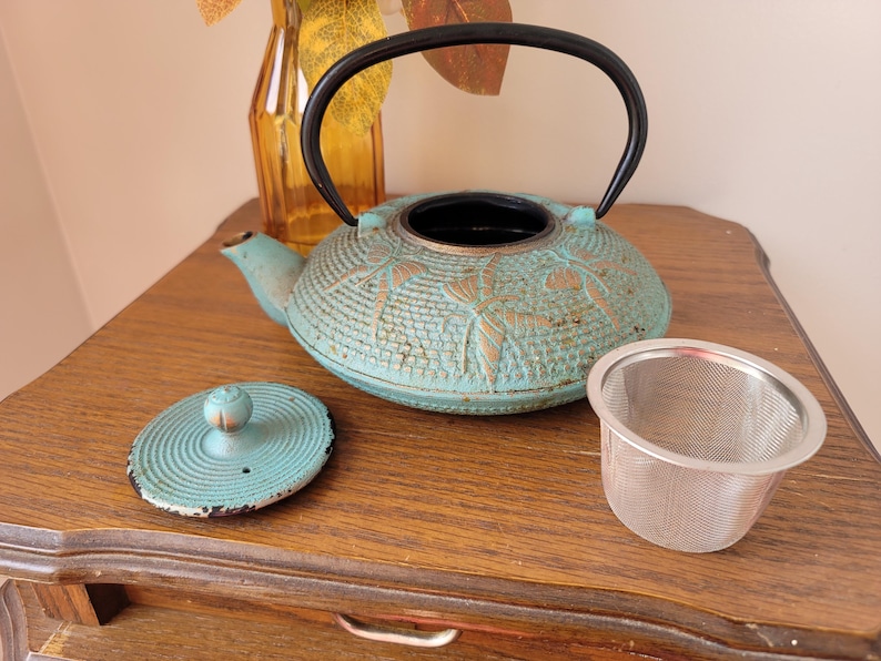 May include: A teal blue cast iron teapot with a black handle and a matching lid. The teapot has a floral design and is sitting on a wooden table with a silver tea strainer.