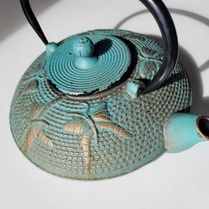 May include: A teal blue cast iron teapot with a gold butterfly design. The teapot has a lid with a round knob and a spout on the side.