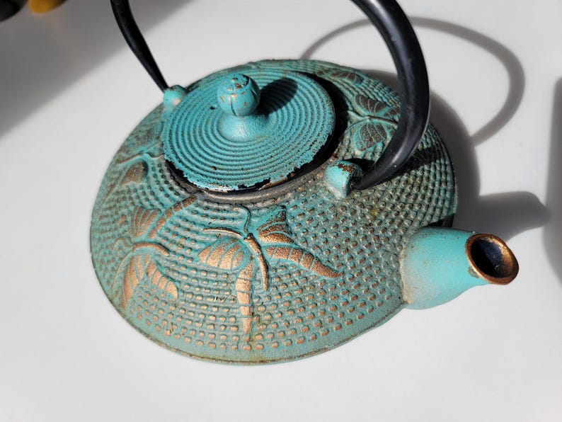 May include: A teal blue cast iron teapot with a gold butterfly design. The teapot has a lid with a round knob and a spout on the side.
