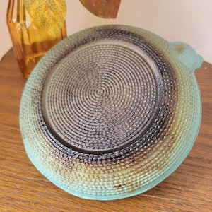 May include: A teal blue ceramic dish with a textured, woven pattern on the bottom. The dish has a slightly raised lip and a small handle on the side.