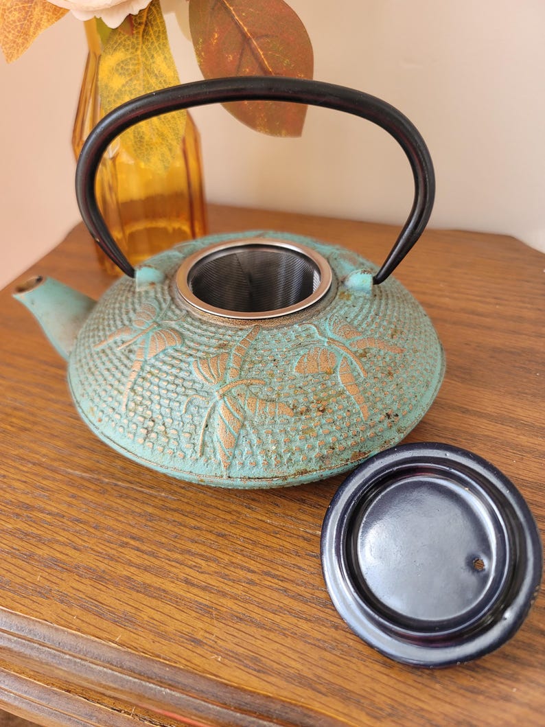 May include: A teal blue cast iron teapot with a gold floral design and a black handle. The teapot has a built-in strainer and comes with a matching lid.