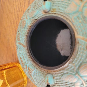 May include: A teal blue ceramic pot with a decorative design of butterflies and floral patterns. The pot has a black interior and a round opening at the top.