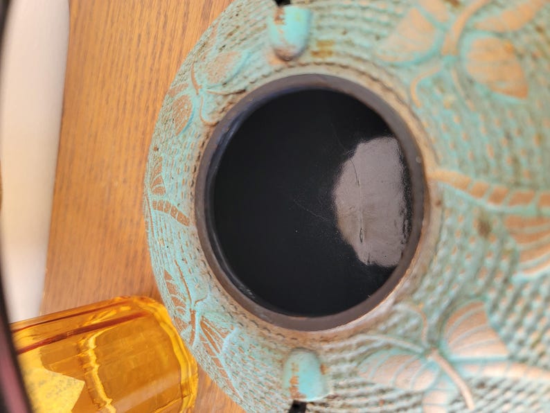 May include: A teal blue ceramic pot with a decorative design of butterflies and floral patterns. The pot has a black interior and a round opening at the top.