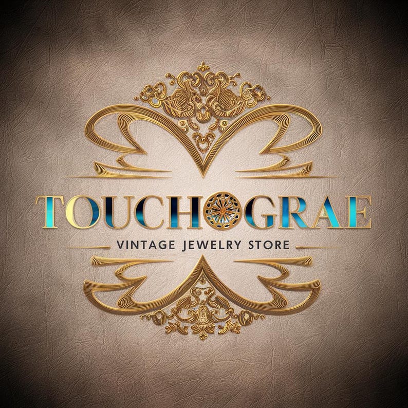 May include: Gold and blue logo for a vintage jewelry store called "Touchograe". The logo features a decorative, ornate design with a circular element in the center.