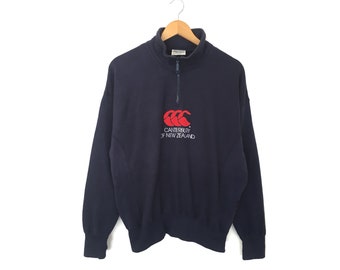 Puede incluir: Una sudadera azul marino con un logotipo rojo y blanco de Canterbury de Nueva Zelanda. La sudadera tiene cuello alto y cremallera.