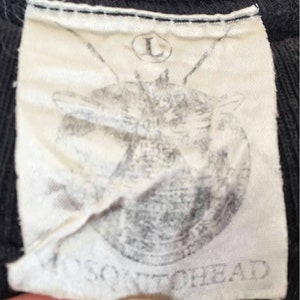 May include: Close-up of a black garment tag with the text "MOSQUITOHEAD" printed in gray. The tag also features a faded graphic and the letter "L" inside a circle, indicating size.