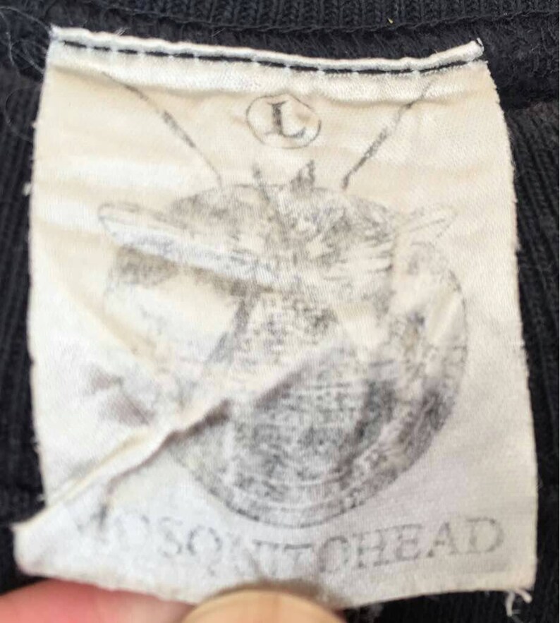 May include: Close-up of a black garment tag with the text "MOSQUITOHEAD" printed in gray. The tag also features a faded graphic and the letter "L" inside a circle, indicating size.