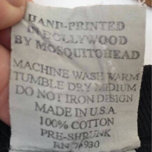 May include: A close-up of a white fabric tag with black text. The tag reads "HAND-PRINTED IN HOLLYWOOD BY MOSQUITOHEAD." Additional text includes washing instructions, "MADE IN U.S.A," "100% COTTON," and "PRE-SHRUNK."