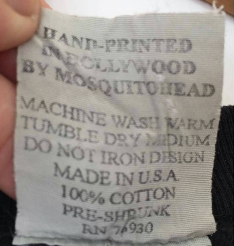 May include: A close-up of a white fabric tag with black text. The tag reads "HAND-PRINTED IN HOLLYWOOD BY MOSQUITOHEAD." Additional text includes washing instructions, "MADE IN U.S.A," "100% COTTON," and "PRE-SHRUNK."