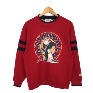 Peut inclure: Sweat-shirt rouge avec un logo bleu et jaune Santa World représentant un Père Noël en dessin animé avec un chien. Le logo comporte le texte "Santa World" dans un cercle avec les mots "Visible to Children and Adults Eyes, Santa Can Be Perceived Through Love, Truth and Imagination" autour du cercle.