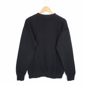 May include: A black sweatshirt hangs from a wooden hanger against a white background. The sweatshirt has long sleeves and a crew neck. The fabric appears to be a soft, comfortable material. The cuffs and hem are ribbed.