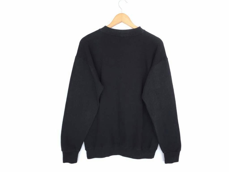May include: A black sweatshirt hangs from a wooden hanger against a white background. The sweatshirt has long sleeves and a crew neck. The fabric appears to be a soft, comfortable material. The cuffs and hem are ribbed.