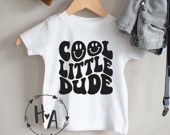 May include: White t-shirt with black text that reads "Cool Little Dude" in a retro font with smiley faces.