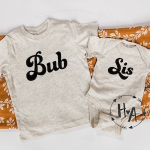 May include: Two beige t-shirts with black retro font. The larger shirt says "Bub" and the smaller shirt says "Lis".