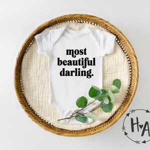 May include: White baby onesie with black text that reads "most beautiful darling." The onesie is lying on a woven basket with a sprig of greenery.