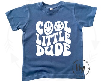 May include: A blue short-sleeved t-shirt with white text that reads "COOL LITTLE DUDE" in a retro font. Two white smiley faces are above the text.
