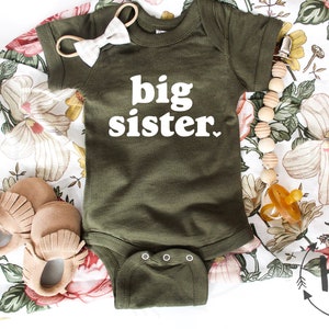 May include: A green baby onesie with the text "big sister" in white. The onesie is lying on a white floral fabric with a pair of brown moccasins, a wooden bead necklace, and a pacifier.