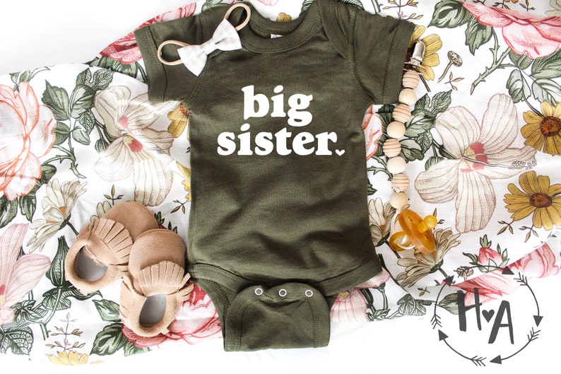 May include: A green baby onesie with the text "big sister" in white. The onesie is lying on a white floral fabric with a pair of brown moccasins, a wooden bead necklace, and a pacifier.
