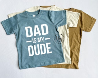 May include: Three t-shirts stacked on top of each other. The top shirt is blue and says "DAD IS MY DUDE" in white letters. The middle shirt is beige and the bottom shirt is brown.