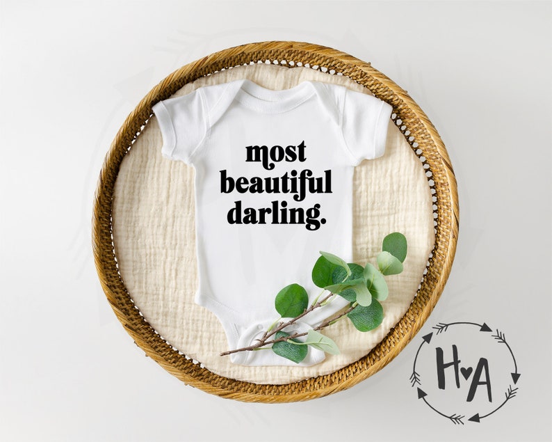 May include: White baby onesie with black text that reads "most beautiful darling." The onesie is lying on a woven basket with a sprig of greenery.