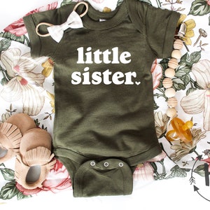 May include: A green baby onesie with the text "little sister" in white. The onesie is on a floral fabric background with a wooden bead necklace, a pair of brown moccasins, and a pacifier.