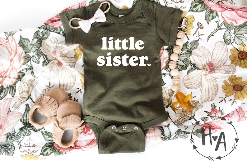 May include: A green baby onesie with the text "little sister" in white. The onesie is on a floral fabric background with a wooden bead necklace, a pair of brown moccasins, and a pacifier.