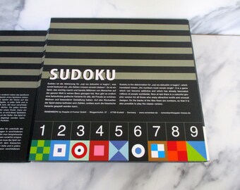 May include: Sudoku game box with a black and white striped background. The box has the word "Sudoku" in large white letters. The back of the box has a description of the game in German and English. The front of the box has a grid with numbers 1 through 9 and colorful symbols.