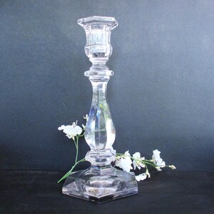 May include: A clear glass candle holder with a hexagonal base and a fluted design. The candle holder is on a black surface with white flowers.