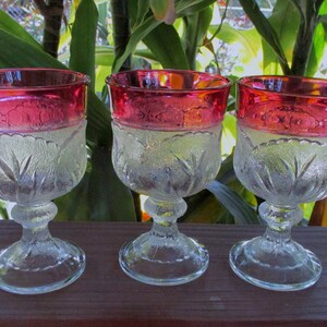 Puede incluir: Tres copas de vidrio transparente con un diseño esmerilado y un borde rojo. Las copas están sobre una superficie de madera.