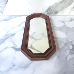 May include: A small, octagonal-shaped mirror with a dark brown wooden frame. The mirror is set against a white marble background.