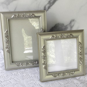 May include: Two silver picture frames with ornate designs. The frames are on a white lace tablecloth.