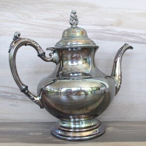 May include: A silver teapot with an ornate handle and lid. The teapot has a round body and a flared base.