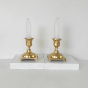 May include: Two gold-coloured candle holders with white candles. The candle holders have a classic design with a flared base and a detailed stem. The candles are white and have a slightly curved shape. The candle holders are placed on a white surface.