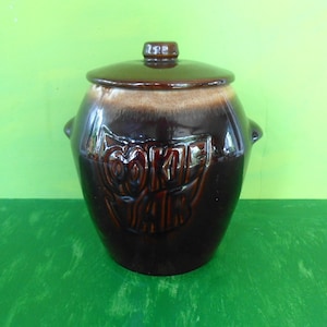 Pode incluir: Jarro de biscoitos de cerâmica marrom envidraçado com tampa. O jarro tem a palavra "COOKIE JAR" em relevo na frente.