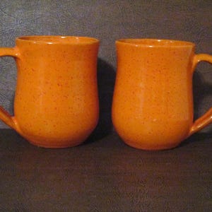 May include: Two orange speckled ceramic mugs with rounded bottoms and handles.