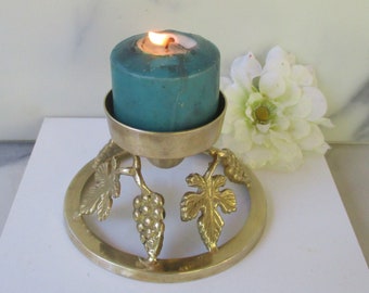 May include: A brass candle holder with a teal candle and a grapevine design. The holder has a round base and three grapevine branches with leaves and grapes.