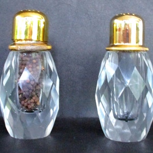 Può includere: Due saliere e pepiere in vetro trasparente con coperchi dorati. Le saliere hanno un design sfaccettato e sono riempite con grani di pepe nero.