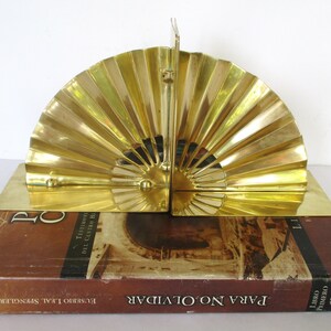 May include: A pair of gold-toned metal bookends shaped like a fan. The bookends are sitting on top of a brown book with the title "Para No Olvidar" in Spanish.
