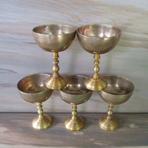 May include: Six vintage brass goblets with a classic design. The goblets have a round bowl shape and are supported by a detailed stem and base. The brass has a warm, aged patina, suggesting they are antique or vintage.