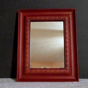 May include: A small, rectangular mirror with a red frame. The frame has a decorative, embossed design around the edge.