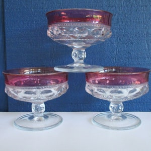 May include: Three clear glass dessert dishes with a ruby red rim. The dishes have a pedestal base and a textured pattern.