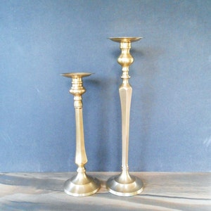 May include: Two gold metal candle holders, one taller than the other, with a simple, elegant design. The candle holders have a round base and a tall, slender stem.