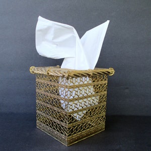 May include: A gold metal tissue box holder with an intricate, ornate design. The holder is square shaped and has a decorative top.