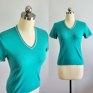 May include: A teal short-sleeved shirt with a white trim around the neckline. The shirt is on a white mannequin.