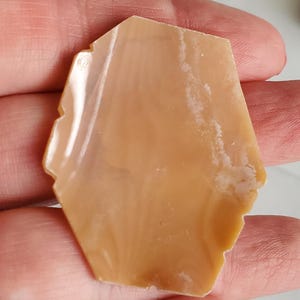 May include: A polished, irregularly shaped stone with a warm, light brown hue. The stone has a smooth, glossy surface with subtle variations in color and texture. The edges are slightly uneven, adding to its natural appearance. The stone is held in a hand.