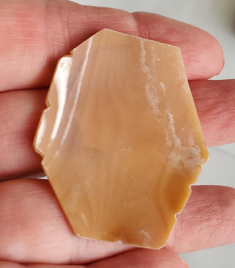 May include: A polished, irregularly shaped stone with a warm, light brown hue. The stone has a smooth, glossy surface with subtle variations in color and texture. The edges are slightly uneven, adding to its natural appearance. The stone is held in a hand.